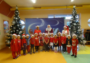 Grupa dzieci stoi z panią dyrektor Marią Królikowską i panią Agnieszką Piekarską, w tle dekoracja świąteczna, choinki.